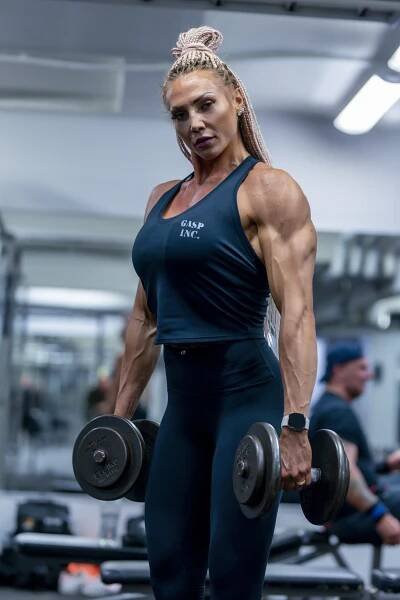 A muscular woman with braided hair lifts dumbbells in a gym, wearing a black tank top and leggings.