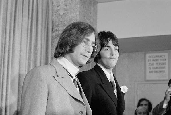 A black and white picture of John Lennon and Paul McCartney standing together. They wear suits.