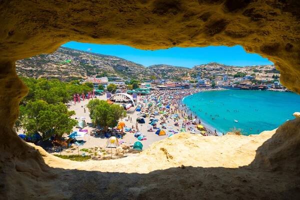 Blick aus einer Höhle auf einen belebten Sandstrand mit bunten Sonnenschirmen. Rechts das blaue Meer, im Hintergrund grüne Hügel und kleine Gebäude. Menschen schwimmen und entspannen am Strand.