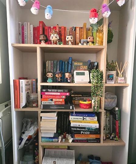 A white bookshelf with Santa decorations hanging across the top, and colorful books, faux plants, and Funko pops on it