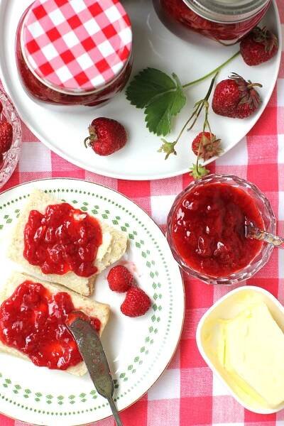 Looking down at the table with a red-checked tablecloth, plate of two biscuit halves spread with low sugar strawberry jam, and butter.