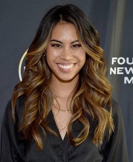 Person smiling at a media event, wearing a stylish dark outfit with waves in their long hair