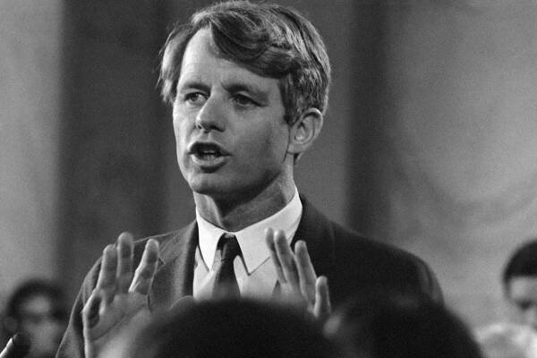 A black and white image of Robert F. Kennedy with his hands raised speaking in public.