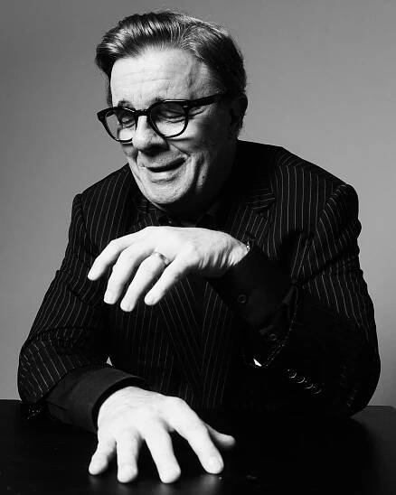 In a black-and-white portrait, a man in a dark pinstriped jacket is closing his eyes and smiling. One hand is on a flat surface, the other is raised in front of him.