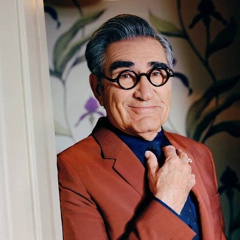 Eugene Levy leans against a wall with his right hand holding the knot of a navy blue tie. He is wearing a dark blue shirt and a rust-colored blazer. He has dark-framed round glasses and dark hair combed back with some gray mixed in. He has one of his distinctive thick eyebrows raised and is smiling with closed lips.