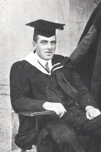 A black-and-white cropped image of a man wearing academic regalia, including a hat and gown. He is sitting in a chair and smiling at the camera. An unknown man's arm is resting on the chair behind him. The image is grainy with age.