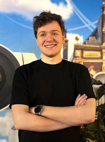 A young man with short curly hair wearing a black t-shirt and a watch, smiling confidently with arms crossed against a colorful background.