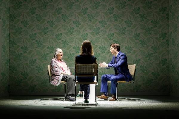 Three people sit at a table, onstage, against a green patterned wall.