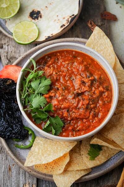 A bowl of chunky salsa roja garnished with cilantro, surrounded by tortilla chips, dried peppers, lime slices, and a partially visible tortilla on a wooden surface.