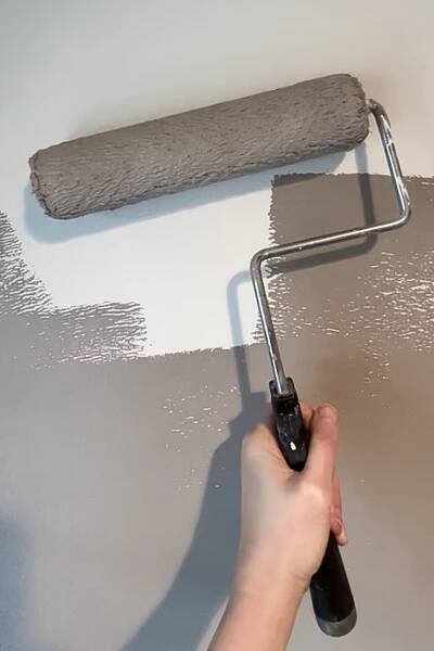 Hand holding a paint roller applying gray paint to a wall.
