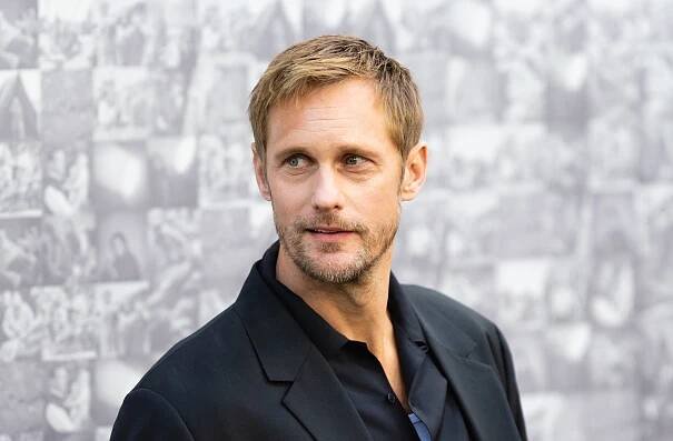 I don't know who this is, but the person is wearing a black suit jacket over a black shirt, standing in front of a backdrop with various black-and-white photos