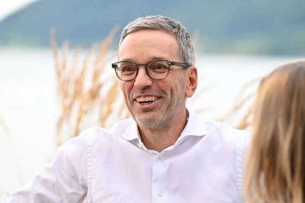 Eine lächelnde Person mit Brille und weissem Hemd steht im Freien mit einem Unschärfehintergrund aus grünem Hügel und goldenem Gras.