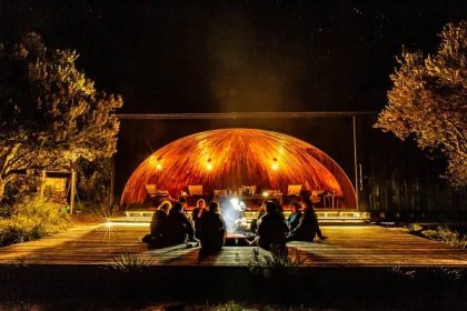 A group of people gather around a fire pit in front of a large, open shelter at night.