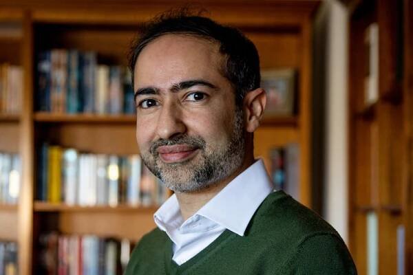 Rahul Khanna, dressed in a button-up shirt and green jumper, stands in front of bookshelves in an office.