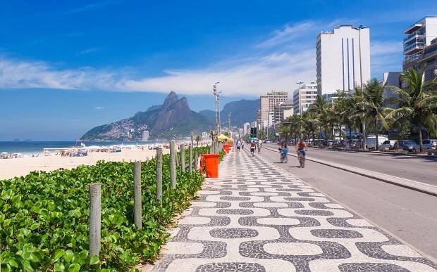 Wide view of Copacabana beachfront promenade in Rio de Janeiro featuring iconic black-and-white wave-patterned mosaic pavement, palm trees, and ocean backdrop.