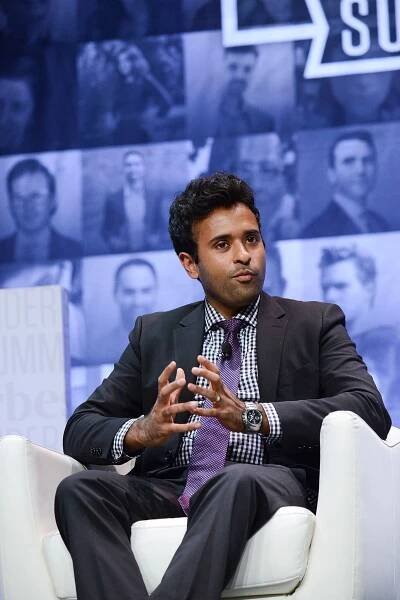 Vivek Ramaswamy sits on a stage and clasps his hands. He is wearing a dark suit and a purple tie. 