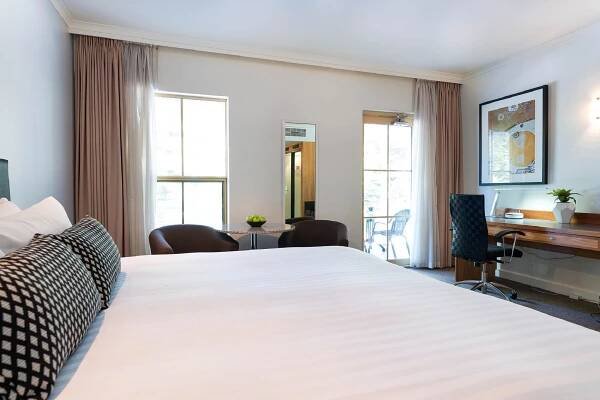 Esplanade Hotel Fremantle - By Rydges Exterior photo The photo shows a well-lit hotel room with a focus on the bed. The bed is large and neatly made with white linens and several pillows, including two decorative black and white patterned pillows in the foreground. 

To the left of the bed, there are t