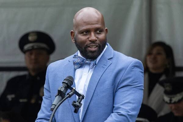 Brian K. Williams, wearing a light blue suit and a bow tie, speaks at a lectern with a microphone.