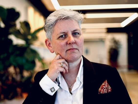 A woman with short grey hair, wearing a dark suit jacket over a white shirt, looks intently into the camera.