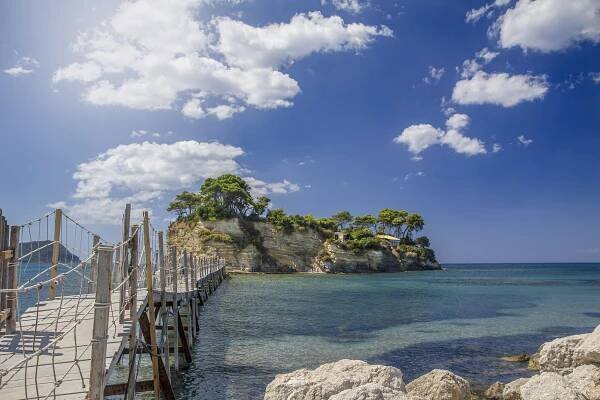 Pláž Laganas, Zakynthos. Most k ostrůvku s vegetací, modré moře a obloha s mraky.