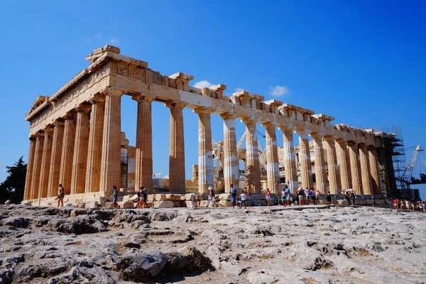 Turisté prozkoumávají Parthenon v Aténách v Řecku pod jasnou modrou oblohou s lešením viditelným za starobylými sloupy.