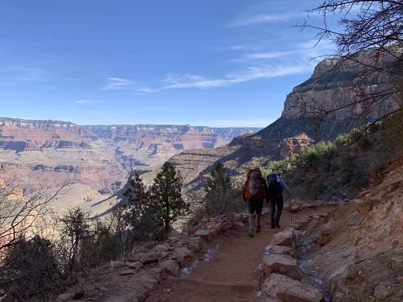 Two backpackers descending Bright Angel Trail, with a view of the Grand Canyon in the background.
