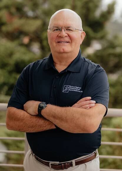 Dr. Ben Gomez in a blue shirt is standing with his arms crossed and smiling.