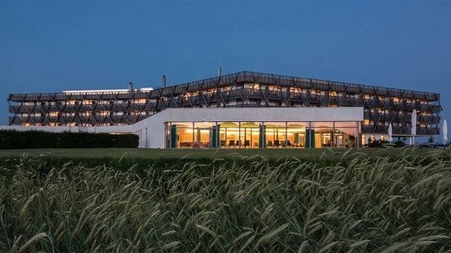 Modernes Hotelgebäude mit erleuchteten Fenstern und umliegenden Feldern bei Dämmerung.