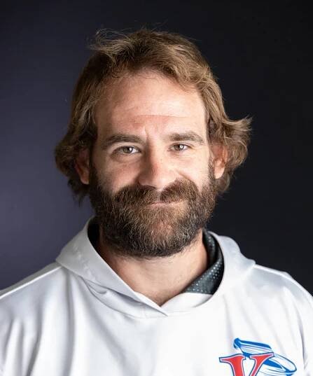 A man with wavy brown hair and a full beard smiles slightly, wearing a white hoodie with a blue and red K logo, standing against a plain dark background.