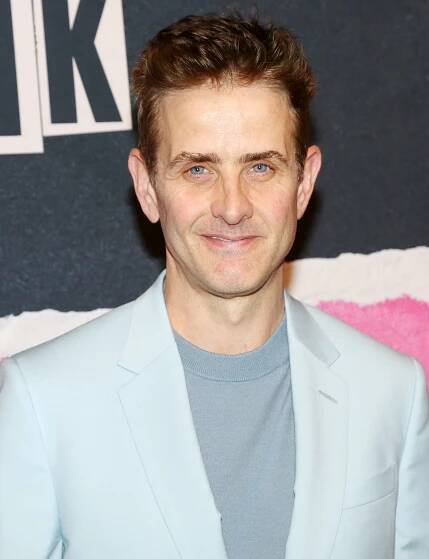 Man in a light suit jacket with a crew-neck shirt at an event, smiling at the camera
