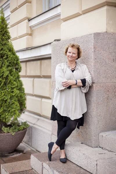 Valokuva hymyilevästä Laura Kolbesta nojaamassa rakennuksen ulkoseinään.