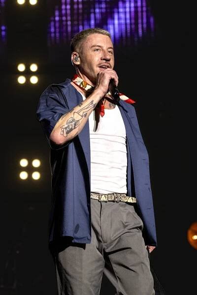 Macklemore onstage in a blue jacket and white T-shirt talking into a microphone