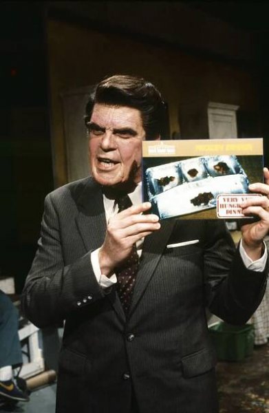 Joe Piscopo as President Ronald Reagan in a suit holding a frozen dinners box, speaking animatedly