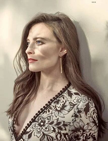 a woman with long brown hair and blue eyes is leaning against a white wall wearing gold earrings