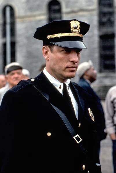 Actor in a police uniform with a serious expression, standing in front of a blurred crowd, possibly a scene from a TV or movie