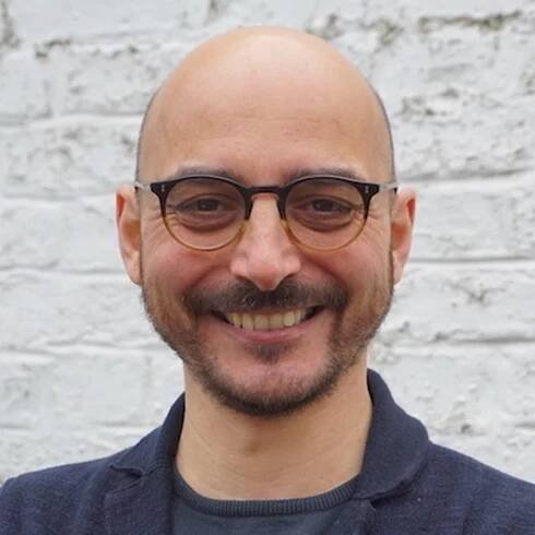 Image of Paolo Lombardi. He has a shaved head, and a short beard and moustache. He's wearing glasses and a gray collarless shirt and blazer.
