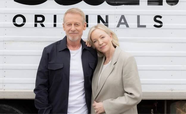 Elderly couple posing in front of a trailer with "ORIGINALS" text behind them, looking content.