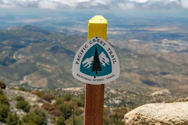 Sign for the Pacific Crest Trail on a wooden post with a picturesque view of rolling mountains, reminiscent of scenes explored during the PCT survey 2018, under a partly cloudy sky.