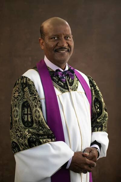 Senior Minister “J” Edgar Boyd stands smiling with his hands clasped over his white church robes with gold trim