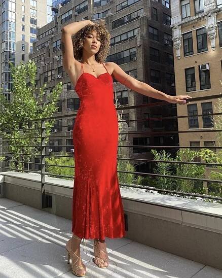 A woman in a red velvet Get The Look: Alexandra Shipp Dress Style dress stands elegantly on a rooftop garden, arms outstretched, against a backdrop of high-rise buildings. She wears gold strappy heels and her