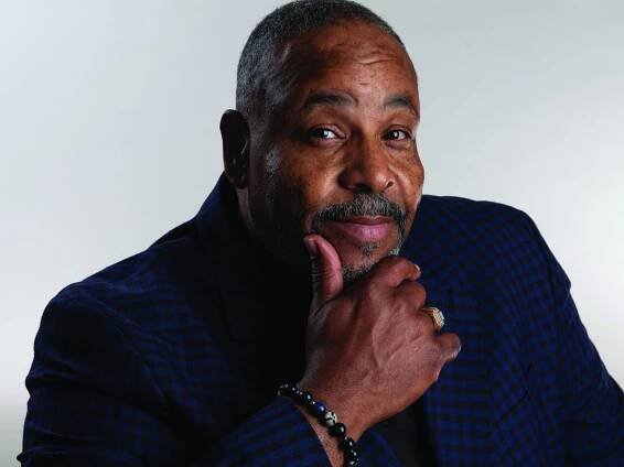 Richard Gibbs gazes at the camera with a friendly, inquisitive look, holding his chin between his thumb and first finger. He wears a dark jacket of blue and black checks and a black beaded bracelet.