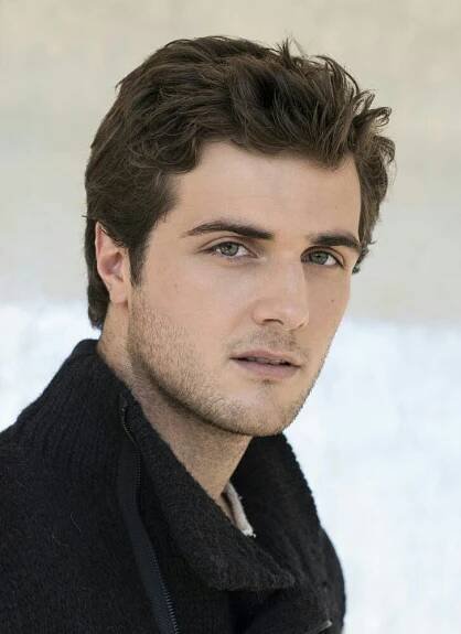 a close up of a person wearing a black jacket and posing for a photo in front of a white wall