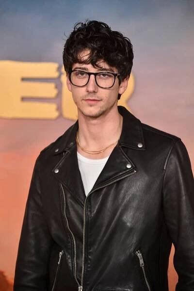 Celebrity in a black leather jacket and glasses at a press event backdrop. The style is casual and relaxed