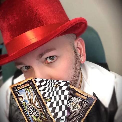 A man wearing a red top hat is holding tarot cards in front of his face