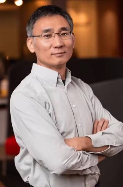 A person in glasses and a light gray button-up shirt stands with their arms crossed, looking confidently at the camera. 