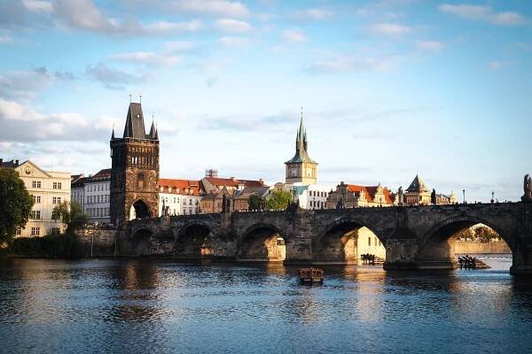 Pohled na Karlův most přes Vltavu v Praze s historickými budovami a kostelními věžemi v pozadí, osvětlený teplým slunečním světlem.