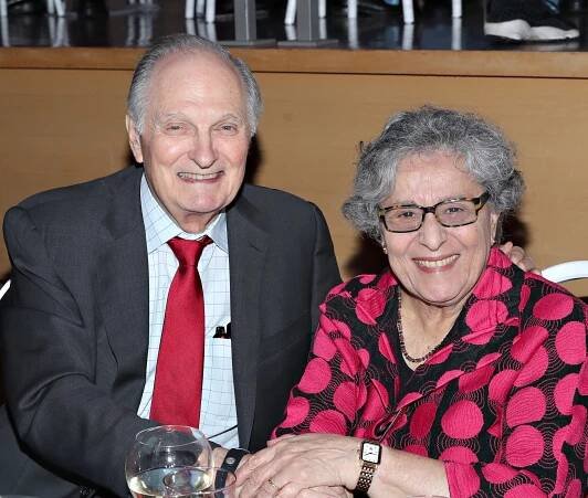 Alan Alda in a suit and tie, sits beside Arlene Alda in a patterned blouse at a table with drinks and decorations, both smiling at the camera