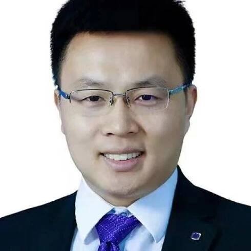 A man with short black hair, wearing glasses, a dark suit, a white shirt, and a purple tie, smiles at the camera against a plain white background.