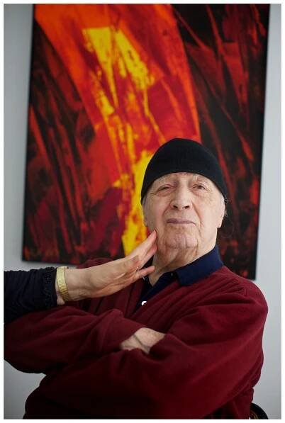 German artist Karl Otto Götz in a red cardigan in front of a red and yellow painting.