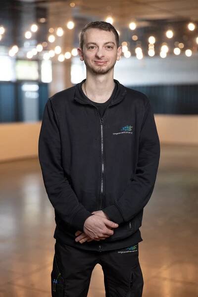 Ein Mann mit kurzen Haaren, einer schwarzen Jacke mit Reißverschluss und einer schwarzen Hose steht mit gefalteten Händen im Innenbereich. Der sanft beleuchtete Hintergrund, dekoriert mit Lichterketten, vermittelt eine warme Atmosphäre – vielleicht bei einem Teamtreffen.